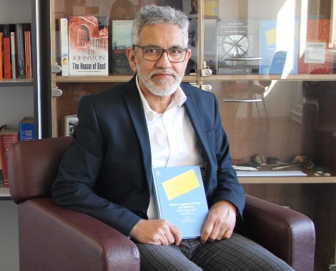 A male wearing a white shirt and navy blue blazer sits in a chair holding a book.